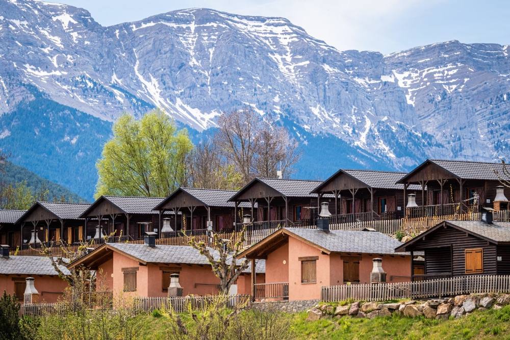 Row of wooden and stone lodges at Cerdanya Viva below a snow-covered mountain range