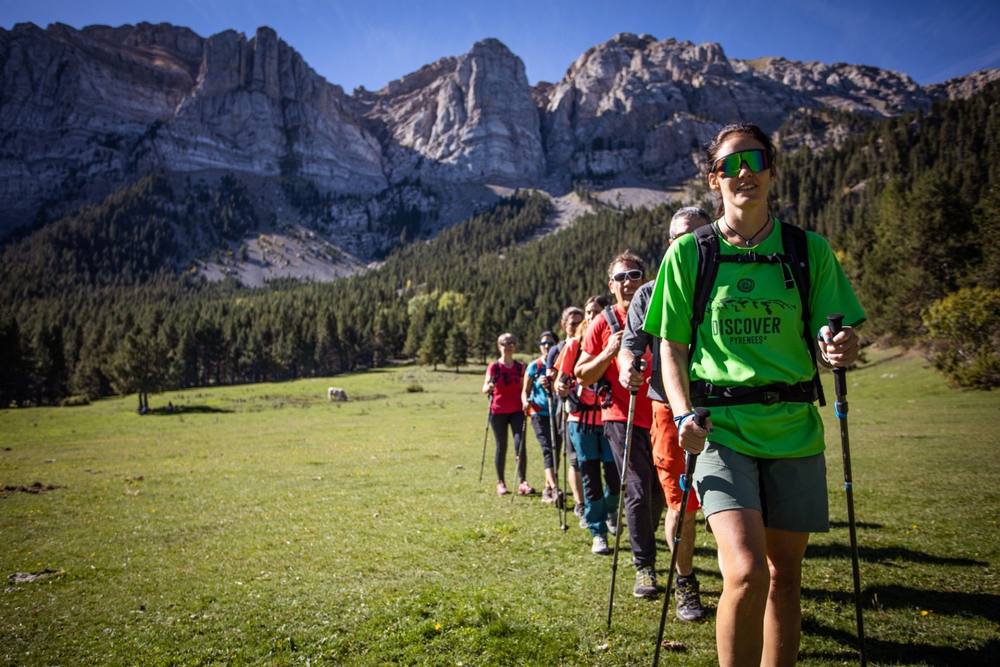 A group walking in single file across a meadow below a Pyrenean rock face